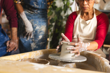 Happy biracial senior female potter with gray hair, using potter's wheel in pottery studio