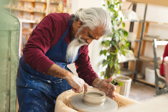 Focused biracial senior male potter working on clay vase using potter's wheel in pottery studio - Powered by Adobe
