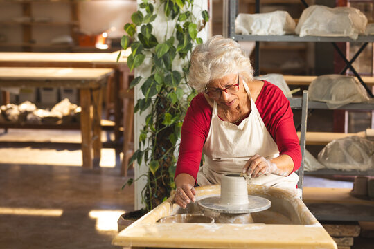 Happy biracial senior female potter with gray hair,using potter's wheel in pottery studio