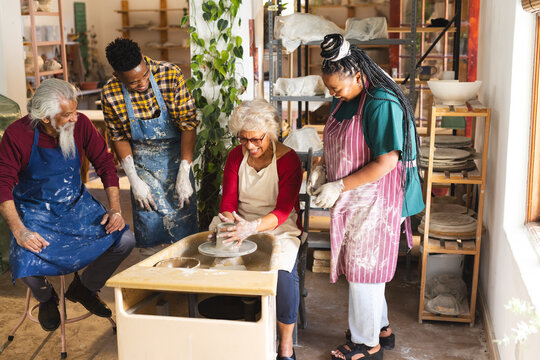 Happy biracial senior female potter with others, using potter's wheel in pottery studio