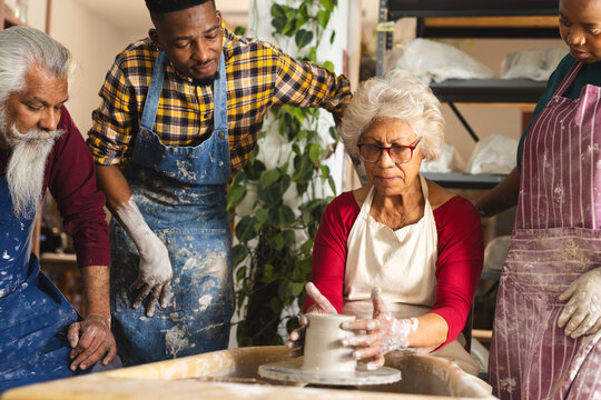 Focused biracial senior female potter with others, using potter's wheel in pottery studio