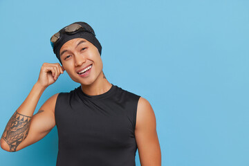 Young asian guy in black top posing isolated over blue background wearing swimming cap with goggles...