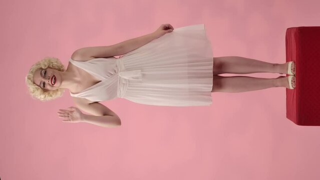Woman in the look of Marilyn Monroe waving her hand in greeting. Woman in white dress and white wig standing full length on pedestal in studio on pink background. Vertical video.
