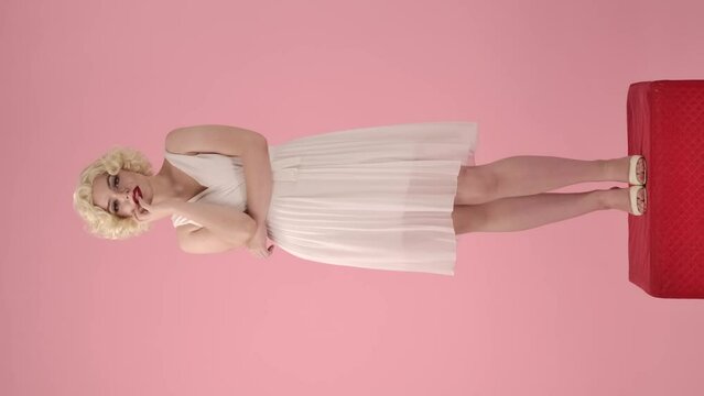 Portrait of a woman smiling sensually and touching her lips coquettishly with her fingers. A woman in the look of Marilyn Monroe standing full length on a red pedestal on a pink. Vertical video.