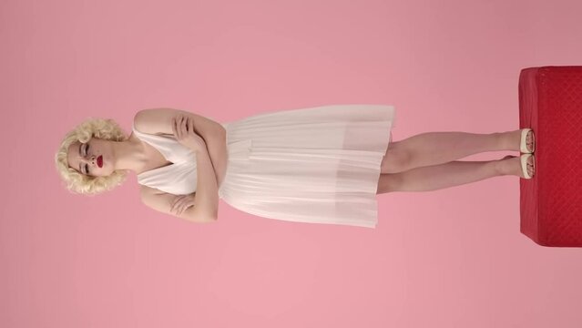 Young woman standing offended, pouting her lips and crossing her arms on her chest. Full length portrait of a woman in the image of Marilyn Monroe on a pedestal in a studio on a pink. Vertical video.