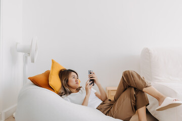 Unhappy Asian Thai woman lying on bean bag sofa, spending time alone at apartment room lonely, holding and using phone waiting for boyfriend texting. get bored and lazy.