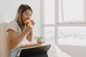 Asian Thai woman having breakfast homemade sandwich on white bed while busy working at home, eating...