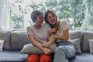 Happy Asian Thai mother and daughter sitting on sofa, smiling and laughing with love, cuddle hugging, good family relationship.