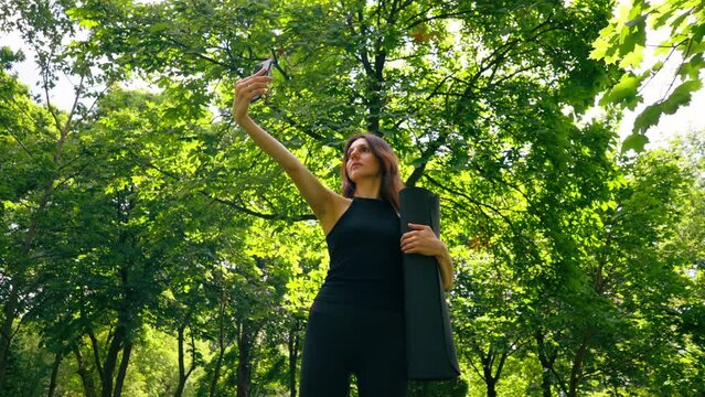 Active beautiful yogi woman during a walk in the park before or after training takes a photo on the phone holds a fitness mat in her hands