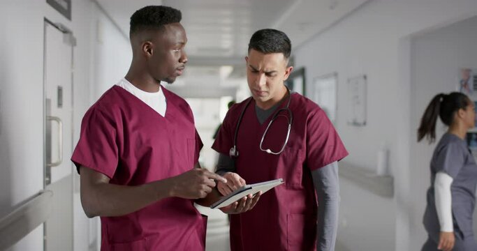 Diverse male doctors discussing work, using tablet in corridor at hospital, slow motion