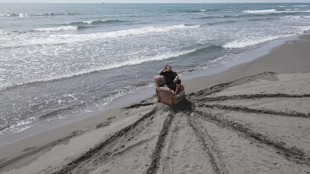 Man pompously squats on old armchair in middle of sandy beach, spreads hair, enjoys power, elements. Artist shoots clip by water, creative entourage Downshifting, psychological retreat, self-knowledge