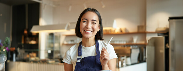Enthusiastic asian female entrepreneur, cafe owner with tablet, wearing shop uniform, standing with tablet and pen and looking excited - obrazy, fototapety, plakaty