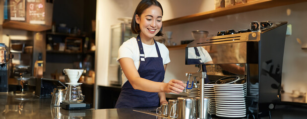 Smiling girl barista in cafe, preparing cappuccino in coffee machine, steaming milk, wearing uniform apron - obrazy, fototapety, plakaty