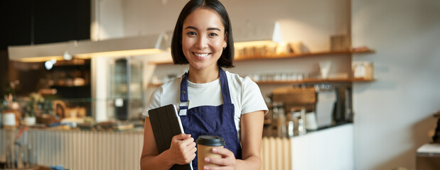 Smiling barista, cafe waitress giving out takeaway coffee order, prepared cappuccino for takeout, standing in blue apron with tablet - obrazy, fototapety, plakaty