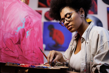 Focused african american artist mixing colors on palette near canvas on easel in studio  - obrazy, fototapety, plakaty