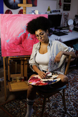 African american artist holding palette and art supplies near painting on easel in workshop