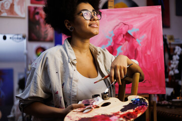 Pleased african american artist holding paint palette and art supplies near canvas in workshop