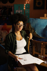 African american artist in eyeglasses holding sketchbook and looking at camera near paintings in...