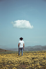 A boy with a white t-shirt and a hat on his head and glasses in his hand with a special and single cloud in the sky in a field full of yellow flowers