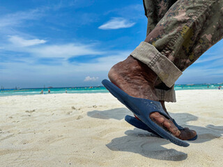 The black feet of an old man wearing blue sandals and military pants walking along the white sandy...