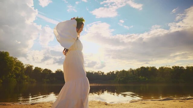 Art portrait happy young woman walks by river bank water lake day blue sky sun light green forest trees back lit. Beauty girl elf enjoy nature. herbal wreath white boho old style dress slow motion