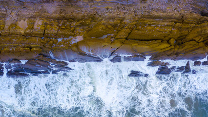 Aerial view of sea and rocks, ocean blue waves crashing on shore. Drone above of turquoise ocean waves crashing and white foaming on huge reef rock stones. Wild sea washing reef rock cliff. 