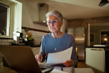 Focused senior woman reviewing a document while using her laptop