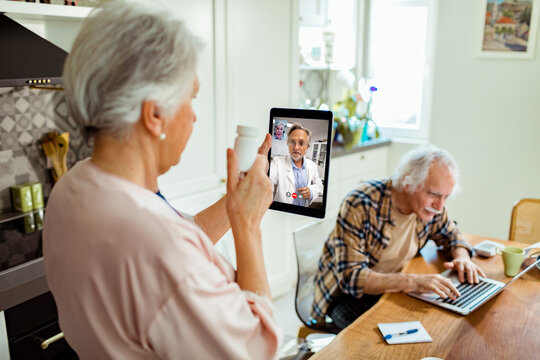 Senior woman discussing medication with her doctor via a telemedicine session while her husband works nearby