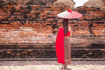 Young woman wearing a red Thai dress and gold accessories stands holding an ancient umbrella with...