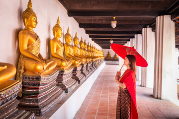 Young woman wearing traditional red Thai dress and golden accessories stands holding a traditional umbrella in the historical site Wat Phutthaisawan Ayutthaya. Thai national costume