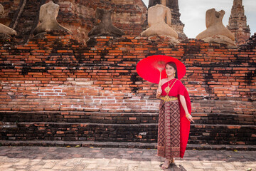 Young woman wearing traditional red Thai dress and golden accessories stands holding a traditional umbrella in the historical site Wat Chaiwatthanaram Ayutthaya. Thai national costume