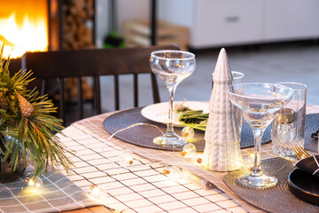 Serving festive table with plates, forks, knives, napkins, glasses close-up near stove fireplace in interior of loft house decorated for Christmas and New Year. Waiting for guests for festive dinner