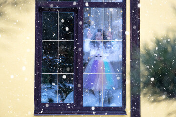 Girl looks out window at snowfall from Cozy festive home outside with the warm light of fairy lights garlands inside - celebrate Christmas and New Year in a warm home. 