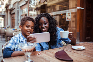 Young African American mother taking selfies with her son in a outdoor cafe sitting area in the city
