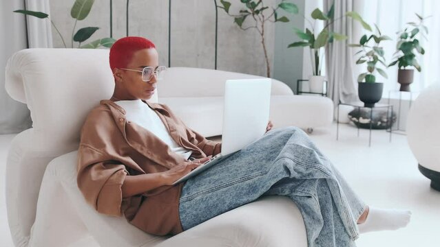 Young positive African American woman working with laptop clutching head and waving hands with smile after noticing stupid mistake sits in comfortable chair in spacious apartment. Freelancer, student