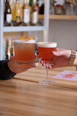 Two alcoholic cocktails in hands over a wooden cafeteria table.