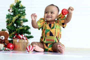 Cute African baby kid in colorful dress sit near decorative Christmas tree and gift box present in white room, happy cheerful little girl child raise hands up, celebrate happy Christmas winter holiday