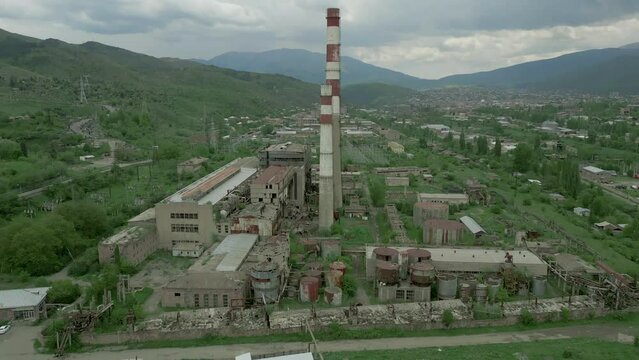 An industrial zone, an old ruined factory, chimneys, buildings with broken windows and trees on the roofs. Around high green mountains in cloudy weather. Drone video. Straight overflight.