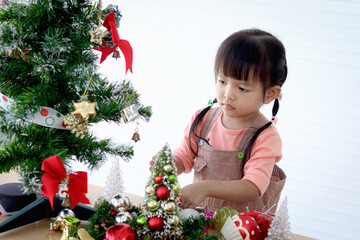 Cute little Asian girl child decorating beautiful Christmas tree with ornaments in white living room. Concentrate kid celebrating winter holiday. Merry Christmas and Happy New year. Happy childhood