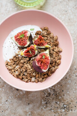 Granola with greek yogurt and torn fig fruits in a roseate bowl, vertical shot on a beige granite background, elevated view