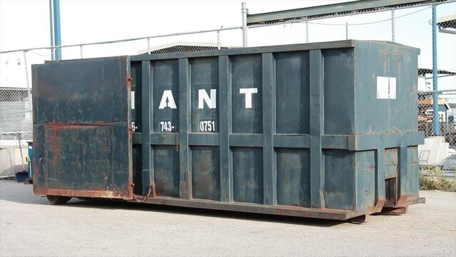 blue container large rental temporary mobile transport dumpster bin with doors open outside in summer on cement pavement in summer with a fence behind