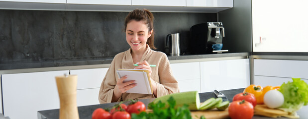 Portrait of beautiful, smiling young woman making list of meals, writing down recipe, sitting in...