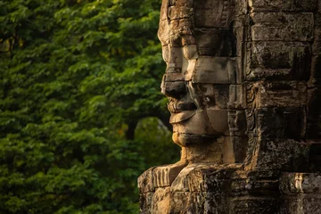 Papier Peint photo Lieu de culte A huge face sculpted in stone, watches over visitors to the temple of Bayon, Angkor, Cambodia, in the background, a wall of green jungle surrounds it