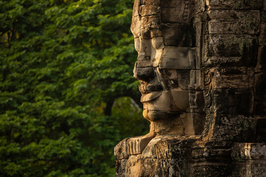 A huge face sculpted in stone, watches over visitors to the temple of Bayon, Angkor, Cambodia, in the background, a wall of green jungle surrounds it