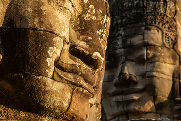 Two stone faces, sculpted at Bayon temple, illuminated by the last rays of evening sunlight,...