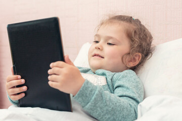 Little girl watching cartoons or playing a game on a tablet in the bedroom at home