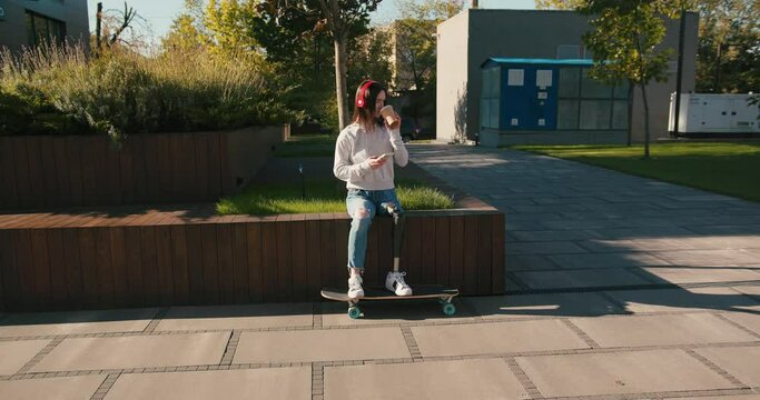 Female with prosthetic leg sitting on longboard. Woman with prosthetic leg wearing red headphones and drinking coffee. Woman with leg prosthesis equipment using smartphone