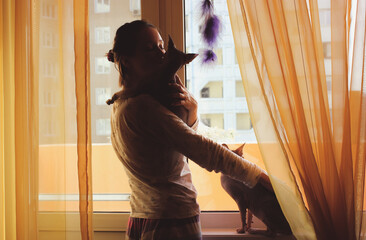A young woman with two lovable cats indoors is looking in a window. Slow living in cozy homy atmosphere. Naked sphynx cat indoors. Sphinx kitties at home. Love, care for pets, animals. Pet sitter. © vita