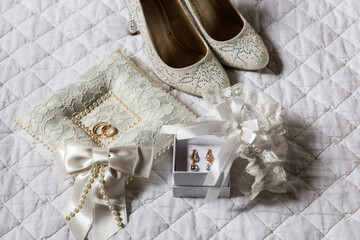 Elegant bridal accessories: jewel-encrusted heels, lace garter, pearl-trimmed ring pillow, and gold earrings on white quilted bedding.
