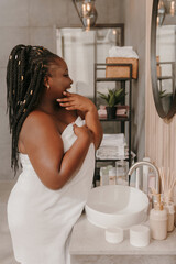 Confident plus size African woman touching face and radiating self-love while looking at the mirror in bathroom